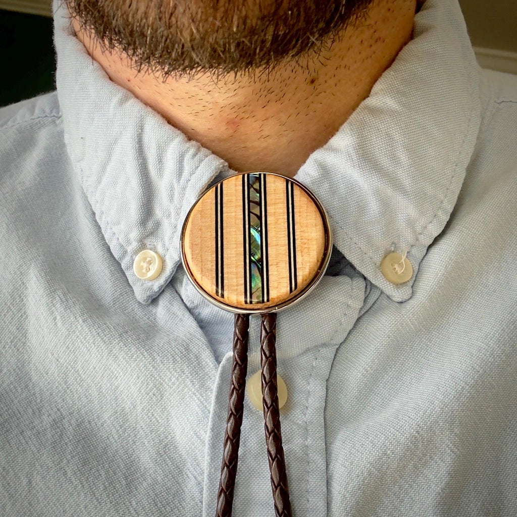 A close-up of a bolo tie worn with a light blue button-down shirt. The wooden slide features vertical black lines and a shimmering abalone inlay. The dark brown braided leather cords hang neatly below.