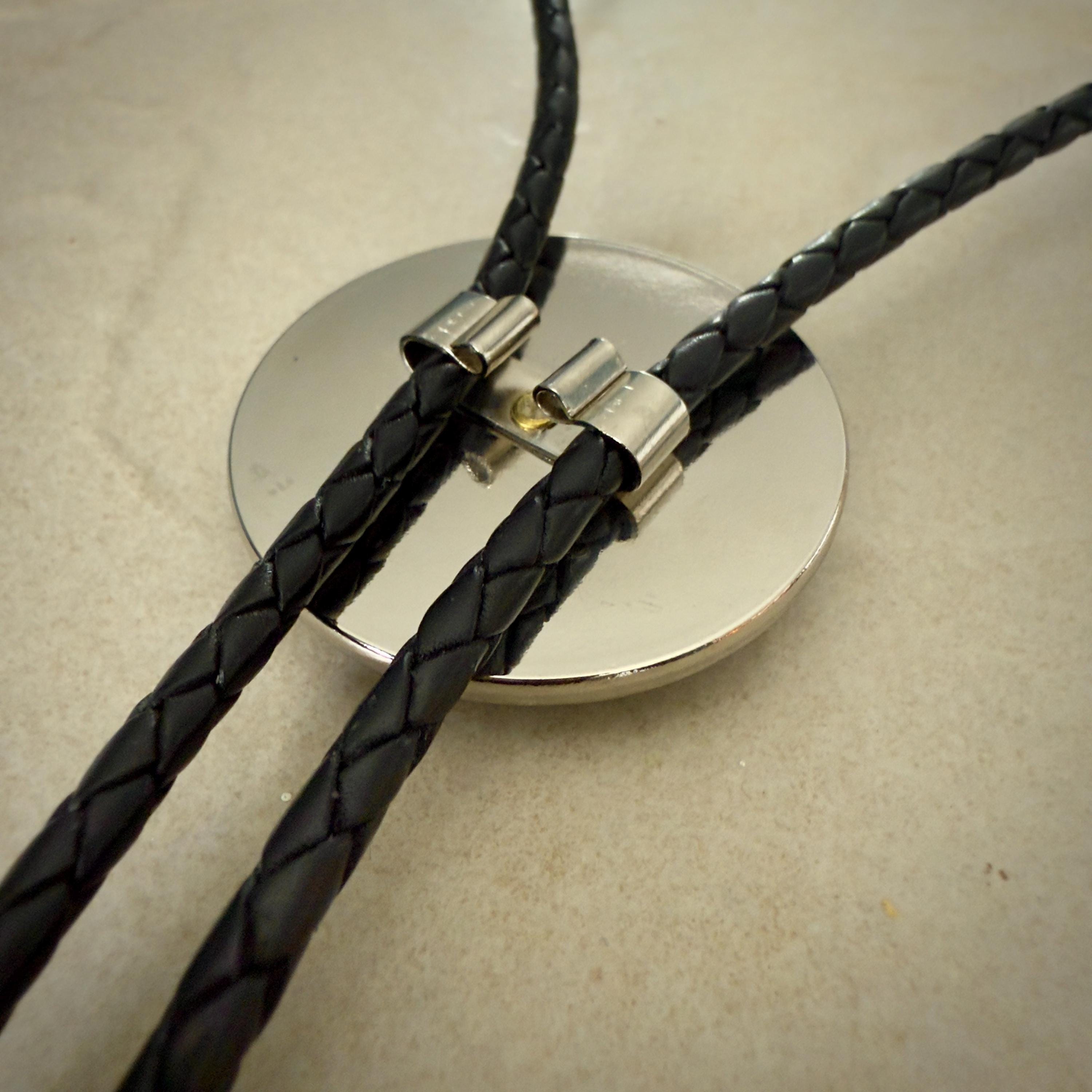 Close-up of a bolo tie resting on a stone surface. The tie features two black braided leather cords held in place by shiny silver metal tips, attached to a circular metallic centerpiece with a smooth, reflective surface.